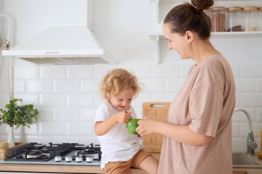 Neşeli sarı dalgalı saçlı küçük kız mutfakta annesiyle yemek yiyor ve tezgahta otururken fincandan nefis bir şeyler atıştırıyor.