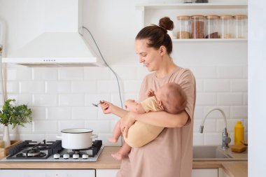 Anne, bebeğini kucağında tutarken mutfakta yemek pişiriyor ve neşeli çocuğuna gülümsüyor. Yemek hazırlıyor ve doğum izni sırasında çocuğuna bakıyor.