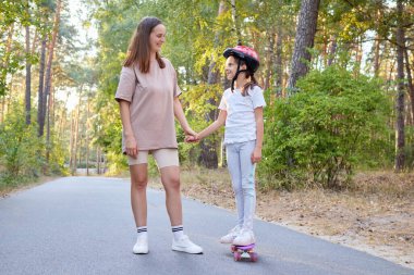 Anne, kaykayla gezen küçük kızı destekliyor. Doğada paten kaymayı öğrenen bir çocuğun yaz etkinliğinin tadını çıkarıyor.