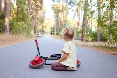 Küçük dalgalı saçlı kız parkta dengeli bisikletinin yanında oturmuş oyunculuk eğitimi sırasında asfalt yolda sürmeyi öğreniyor.