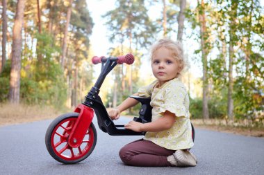 Şirin sarı dalgalı saçlı kız çocuğu denge bisikletiyle dışarıda çalışıyor yaz eğlenceleri için yeni bisikletini çalışıyor.