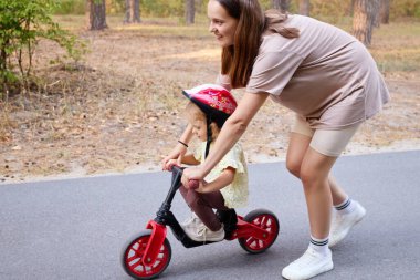 Gülümseyen genç bir anne, dengeli bisiklette çocuğuna destek oluyor ve ona eğlenceli yaz günlerinde parkta sürmeyi öğretiyor.