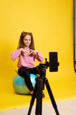 Neşeli çocuk spor yaparken spor yapıyor. Fitness topuyla. Elleriyle kalp şekli oluşturuyor. Aynı zamanda spor sitesinde akıllı telefon kullanarak spor yapıyor.