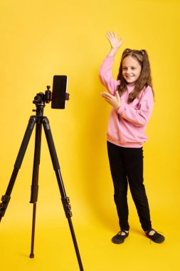 Küçük neşeli pozitif çocuk, internet kanalı içeriği için cep telefonuyla müzik videosuna geçiyor. Çocukluğun ve teknolojinin sarı arka planda izole edilmesinin keyfini sürüyor.