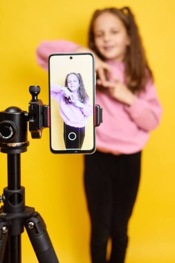Gülümseyen küçük kız çocuğu dans performansını akıllı telefon ve tripodla kaydediyor gülümseyerek kalp şeklindeki aşk jestini gösteriyor yaratıcılığını ve coşkusunu online izleyicilerle paylaşıyor.