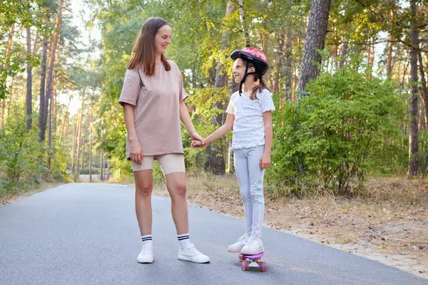 Anne, kaykayla gezen küçük kızı destekliyor. Doğada paten kaymayı öğrenen bir çocuğun yaz etkinliğinin tadını çıkarıyor.