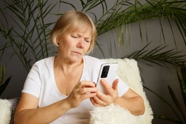 Evde akıllı telefonunu tutan üzgün olgun bir kadın interneti karıştırıyor üzgün ve hoşnutsuz görünüyor cep telefonunda dijital teknolojiyle sosyal medyada sohbet ediyor.