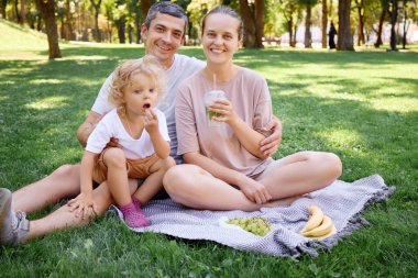 Mutlu genç baba, anne ve küçük kız sıcak güneşli bir günde çimenlerde oturup mutlu bir şekilde ailece dinlenip eğleniyorlar.