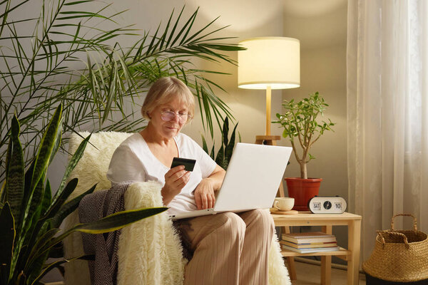 Mature woman senior customer at home using her laptop for online shopping holding credit card to complete her payment and make purchase enjoying convenience of internet banking