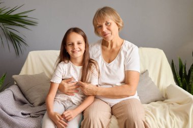 Mutlu son sınıf anneannesi mutlu torununu kucaklıyor. Kanepede rahatça oturuyor. Küçük kız gülümsüyor ve büyükannesini ziyaret ediyor.
