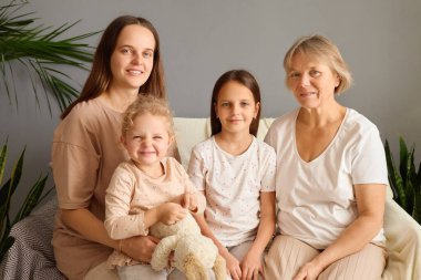 Beyaz bir kadın bebeğini kucağında yaşlı annesi ve büyük kızıyla koltukta otururken sıcak bir evde gülücükler saçarak ailece eğleniyor.