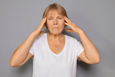 Hüsrana uğramış olgun bir kadın, beyaz tişört giyiyor, baş ağrısı ve migrenden dolayı yüzünü buruşturuyor, hasta ve rahatsız hissediyor. Gri arka planda tek başına ayakta durmaktan rahatsız oluyor.