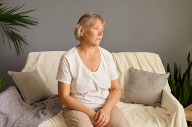 Oturma odasındaki koltukta oturan düşünceli olgun bir kadın uzaktaki hayata bakıyor düşünceli, yaşlı bir kadın evdeki kanepede dinleniyor ve melankolik hissediyor.