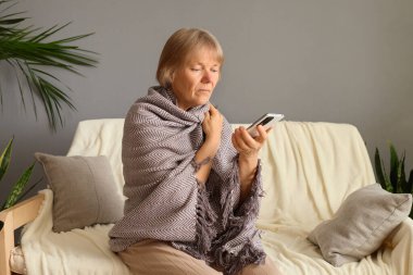 Ekoseli battaniyeye sarılmış olgun bir kadın evinde kanepeye oturmuş akıllı telefondan alışveriş uygulamalarını izliyor sosyal ağda teknoloji haberleri okumaktan keyif alıyor.