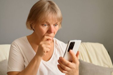 Odaklanmış olgun bir kadın internet üzerinden akıllı telefonun odaklanması ve ciddi olması üzerine sosyal medya uygulamaları kullanarak evdeki koltukta oturan bağlantıyı kontrol ediyor.