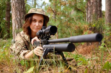 Kamuflaj teçhizatıyla odaklanmış bir nişancı, taktik eğitim sırasında ağaçlık bir alanda dürbüne nişan alarak yere kondu..
