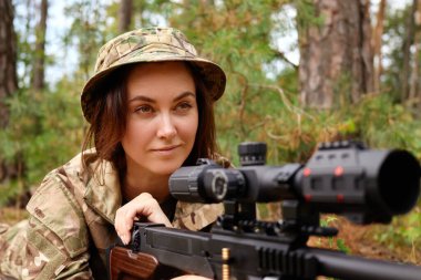 Kamuflaj ve şapka giyen bir asker, sık bir ormanda tüfeğini doğrultuyor. Eğitimdeki bir sonraki hamlesine hazırlanırken ifadesi yoğun bir odaklanma ve kararlılık gösteriyor..