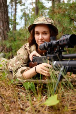 Kamuflaj kıyafeti giymiş bir asker, eğitim sırasında konsantrasyonunu sağlayan keskin nişancı tüfeğini ormana yerleştiriyor..