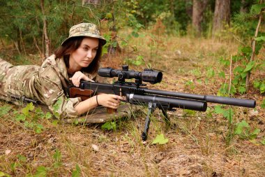 Askeri kamuflajlı bir kişi, bir keskin nişancı tüfeğini çimen ve ağaçlarla dolu doğal bir ortamda nişan almaya odaklanır. Eğitim sırasında taktiksel hassasiyet gösterir..