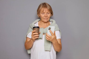 Senior woman drinking coffee chatting on her mobile addicted to social media reading news searching online browsing web page doing shopping in web store standing isolated over background clipart