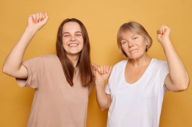 Genç bir kadın ve yaşlı bir kadın kutlama için sevinçle kollarını kaldırıyor, her ikisi de geniş bir gülümsemeyle, güneşli sarı bir arka plana karşı canlı bir mutluluk ortamı yaratıyorlar..