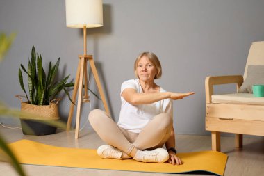Sıcak ve davetkar bir oturma odasında, son sınıf öğrencisi bir kadın sarı bir yoga minderinde oturur, sakin bir ortamda sakin bir sabah geçirirken esneme egzersizleri yapar..