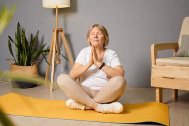 Son sınıftaki bir kadın yoga minderinde bağdaş kurarak oturur, elleri dua pozisyonunda, bitkiler ve doğal ışıkla dolu, kapalı bir ortamda sükuneti ve farkındalığı kucaklar..