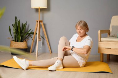Modern bir oturma odasında, rahat kıyafetler giymiş son sınıf bir kadın yoga yaparken saatini kontrol eder, denge ve fitness rutininde farkındalığı ön plana çıkarır..