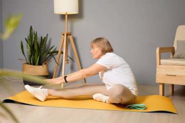 Kadın yoga minderinin üzerinde oturuyor, esneklik egzersizi yaparken ayaklarına doğru uzanıyor. Kapalı alandaki bitkiler rahat mekana yeni bir hava katar..