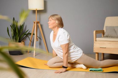 Son sınıf öğrencisi bir kadın oturma odasındaki sarı bir paspasa yoga yaparken esnekliğini ve farkındalığını arttırıyor. Güneş ışığı uzayı aydınlatır, barışçıl bir atmosfer yaratır..