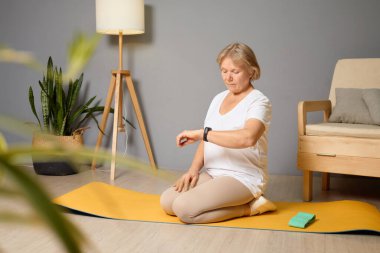 Son sınıf öğrencisi bir kadın sarı bir yoga minderinde bacak bacak üstüne atmış halde oturuyor, akıllı hareketlerini kontrol ediyor ve bitki ve doğal ışıkla dolu modern oturma odasında huzur içinde meditasyon yapıyor..