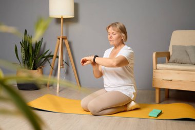 Olgun bir kadın oturma odasındaki sarı yoga minderinde oturur ve meditasyon yapar. Odada bir lamba, bitki ve basit mobilyalar var..