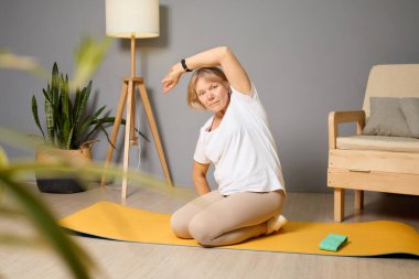 60 'lı yaşlarda bir kadın sarı bir yoga minderinin üzerinde esneme egzersizi yapıyor. Etrafı bitkiler ve doğal ışıkla çevrili basit ama davetkar bir oturma odası ile çevrili..