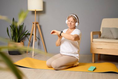 Son sınıf öğrencisi bir kadın, oturma odasındaki sarı bir paspasın üzerinde yoga yapıyor, kulaklık takıyor ve spor ayakkabılarını ya da müzik kontrolünü kontrol ediyor..