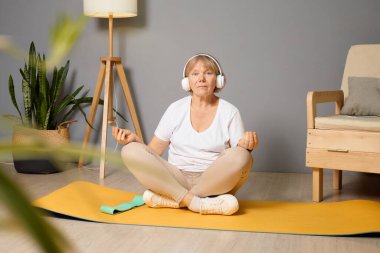 Son sınıf öğrencisi bir kadın evdeki yoga minderinde bağdaş kurup kulaklıkla müzik dinliyor ve sakin, bitki dolu bir ortamda farkındalık yaratıyor..