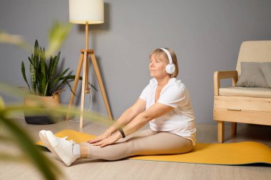 Yaşlı bir kadın yoga minderinin üzerine nazikçe uzanır, kulaklık takar, parlak ve davetkar bir oturma odasında bitkilerle ve rahat bir sandalyeyle..
