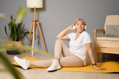 Son sınıf öğrencisi bir kadın, iyi aydınlatılmış bir oturma odasında sarı yoga minderinde oturur, kulaklık takar ve ev egzersizi yaparken müziğinin keyfini çıkarır..