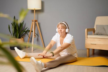 Bir kadın yoga minderinde oturuyor, kulaklık takarak bacaklarını esnetiyor. Parlak oturma odasında bir lamba ve rahat bir atmosfer yaratan ahşap bir sandalye var..