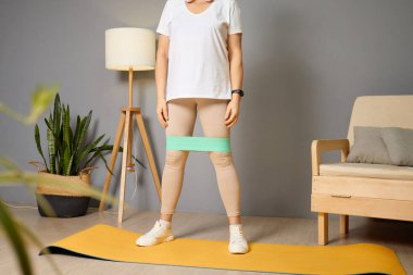Kadın yoga minderinin üzerinde duruyor, bacaklarının etrafına bir direniş bandı takıyor, iyi ışıklandırılmış ve eve yeşillikle dolu bir spor salonunda güç antrenmanına odaklanıyor..