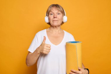 A woman wearing headphones expresses enthusiasm with a thumbs up gesture, holding a rolled yoga mat, set against a vibrant yellow backdrop. It conveys positivity and readiness for practice. clipart