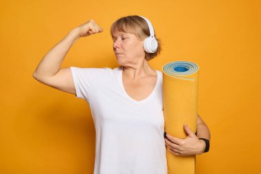 Kendinden emin, kulaklık takan yaşlı bir kadın, yoga minderini tutarken pazılarını esnetiyor. Parlak sarı arka plan, onun fitness coşkusuna enerji katıyor, aktif yaşam tarzını gösteriyor..