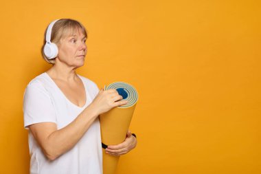 An older adult stands against a vibrant yellow backdrop, holding a rolled yoga mat. She wears headphones, ready for a yoga practice, showcasing a focus on wellness. clipart