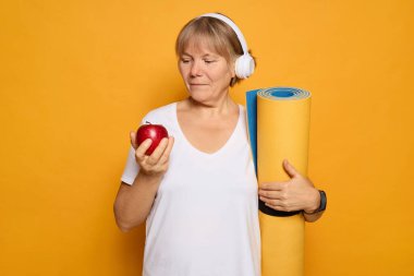 Kulaklık takan bir kadın bir elinde sarı yoga minderi tutarken bir elinde kırmızı bir elmayı inceliyor. Turuncu bir arka planda duruyor, antrenmana hazır..