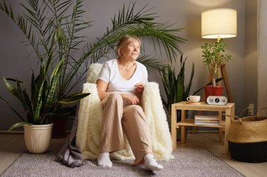 Yaşlı bir kadın, yeşil ve yumuşak ışıklandırma ile çevrili rahat bir sandalyeye rahatça oturur. Düşünceli görünüyor, şık yaşam alanında huzurlu bir anın tadını çıkarıyor..