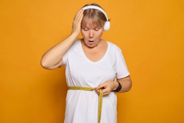 A woman stands confidently against a vibrant yellow backdrop, wearing headphones and measuring her waist with a tape measure while expressing surprise at the result. clipart