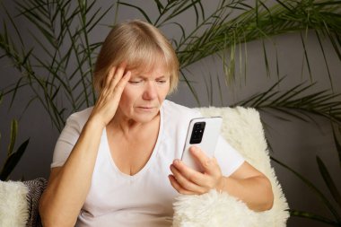 Olgun bir kadın, ev bitkileriyle çevrili yumuşak bir sandalyeye rahatça oturmuş akıllı telefonuyla kitap okurken endişeli görünüyor. Atmosfer sakin ve sakin..