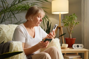 Konforlu, aydınlık bir oturma odasında yaşlı bir kadın elinde kredi kartıyla akıllı telefonunu dikkatle inceliyor. Rahatlatıcı bir çevrede bitkiler ve yumuşak mobilyalar bulunur..