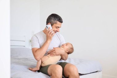 Bir baba yatakta oturuyor, telefonda konuşurken gülümsüyor ve bebeğini kucaklıyor. Parlak, basit yatak odası ebeveynliğin besleyici bir anını yakalıyor..