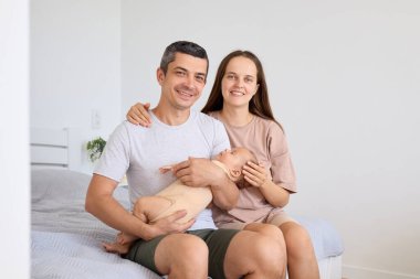 Bir çift, yeni doğan bebeklerini huzurlu bir yatak odasında tutarken sıcak bir şekilde gülümsüyor. Yumuşak ışıklandırma ve basit dekor mutluluk ve sevgiyle dolu huzurlu bir atmosfer yaratır..