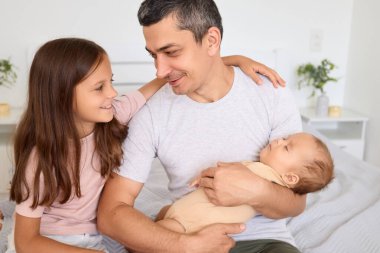 Bir baba, yeni doğmuş bebeğini sıcak ve davetkar bir odada kucağında tutarken kızına gülümser..